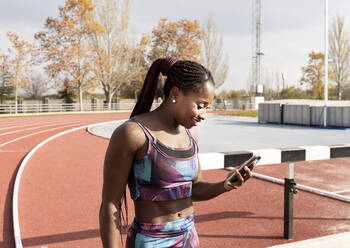 Lächelnde Sportlerin mit geflochtenen Haaren, die an einem sonnigen Tag ihr Smartphone auf der Rennstrecke benutzt - JCCMF00888