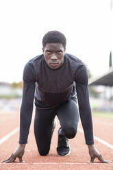 Confident male athlete kneeling on starting line of track against clear sky - JCCMF00899