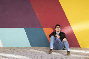 Smiling young man with eyes closed closed listening music while sitting against colorful wall - IFRF00300