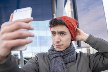 Junger Mann, der sein Strickzeug anpasst, während er ein Selfie mit seinem Smartphone in der Stadt macht - IFRF00313