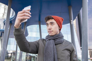 Junger Mann mit Schal nimmt Selfie gegen moderne Gebäude in der Stadt - IFRF00314