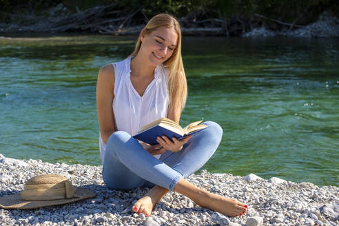 Lächelnde blonde Frau sitzt im Schneidersitz und liest ein Buch am Fluss - LBF03313