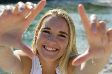 Fröhliche junge Frau zeigt Fingerrahmen, während sie am Seeufer an einem sonnigen Tag steht - LBF03315