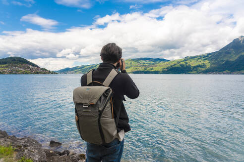 Männlicher Tourist mit Rucksack fotografiert Sarner See gegen bewölkten Himmel in der Schweiz - TAMF02754