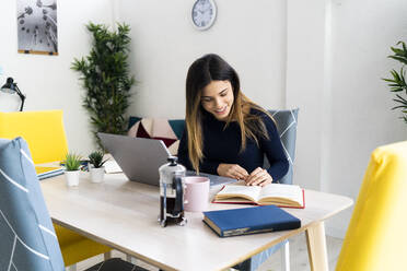 Smiling student with laptop reading book while sitting on table against white wall at home - GIOF10607