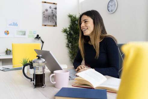 Happy young female freelance worker with laptop and book looking away at home - GIOF10610