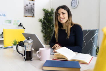 Smiling female freelancer with laptop and book sitting on chair in living room - GIOF10611