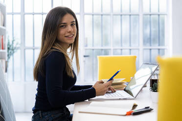 Smiling student with laptop holding mobile phone while sitting on table at home - GIOF10616