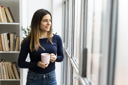 Lächelnde Freiberuflerin mit Kaffeetasse, die durch das Fenster schaut, während sie zu Hause steht - GIOF10620