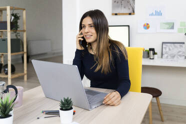 Lächelnde Freiberuflerin beim Telefonieren, während sie zu Hause am Laptop sitzt - GIOF10626