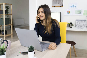 Smiling female freelancer on phone call while sitting by laptop at home - GIOF10626