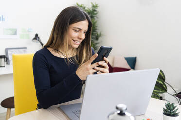 Smiling freelancer worker using mobile phone while sitting by laptop in living room - GIOF10628