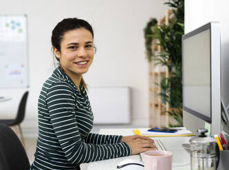 Happy female professional sitting on desk by computer at work place - GIOF10631