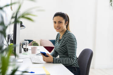 Lächelnde kreative weibliche Fachkraft mit Smartphone sitzt am Schreibtisch neben dem Computer im Büro - GIOF10634