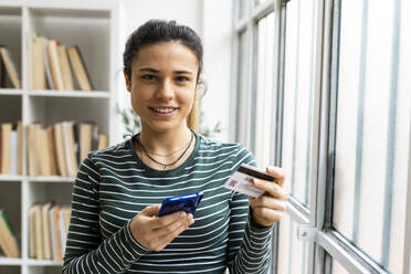 Lächelnde junge Frau mit Smartphone und Kreditkarte, die am Fenster ihres Arbeitsplatzes steht - GIOF10641