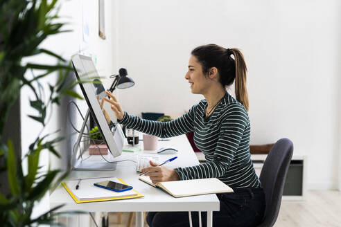 Lächelnde junge Geschäftsfrau berührt den Bildschirm eines Computers am Arbeitsplatz - GIOF10643