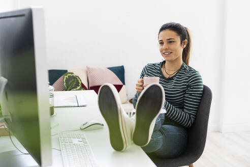 Lächelnde weibliche Fachkraft mit Kaffeetasse, die auf den Computer schaut, während sie die Beine auf dem Schreibtisch im Büro abstützt - GIOF10644