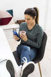 Female entrepreneur using smart phone while holding coffee cup at work place - GIOF10645