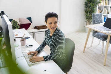 Lächelnde junge Geschäftsfrau, die wegschaut, während sie am Schreibtisch vor einem Computer im Büro sitzt - GIOF10646