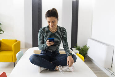 Junge Unternehmerin, die ein Mobiltelefon benutzt, während sie auf einem Tisch im Büro sitzt - GIOF10658