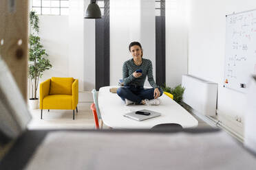 Lächelnde weibliche Fachkraft, die ein Smartphone hält, während sie auf einem Tisch im Büro sitzt - GIOF10660