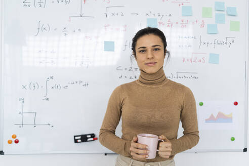 Eine Studentin hält eine Kaffeetasse in der Hand, während sie zu Hause am Whiteboard steht - GIOF10666