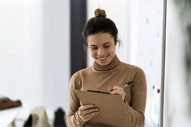 Happy young female student writing on clipboard while studying in living room - GIOF10678