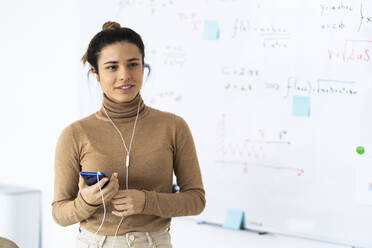 Eine Studentin hält ihr Handy in der Hand, während sie zu Hause über Kopfhörer Musik hört - GIOF10682