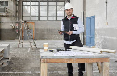 Lächelnder männlicher Architekt mit digitalem Tablet an einem Tisch auf einer Baustelle - VEGF03542