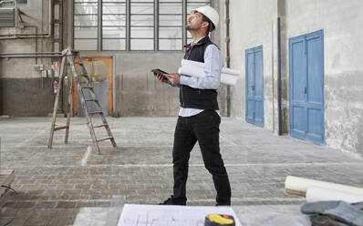 Male architect with digital tablet looking up while standing in constructing building - VEGF03543