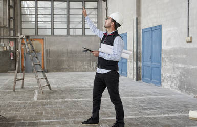 Male architect with digital tablet pointing while looking up in constructing building - VEGF03544