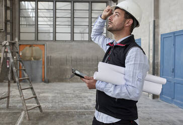 Male architect with digital tablet and blueprints looking up while standing in building - VEGF03545