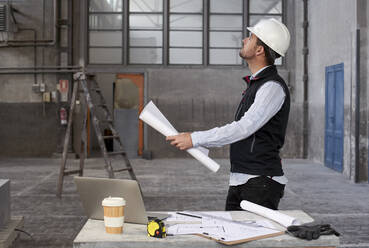 Männlicher Architekt, der einen Bauplan hält und nach oben schaut, während er in einem Gebäude steht - VEGF03549