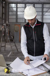 Male architect analyzing blueprint on table while standing inside construction site - VEGF03551