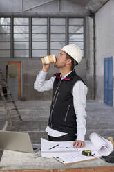 Männlicher Architekt trinkt Kaffee, während er an einem Tisch in einem Gebäude steht - VEGF03563
