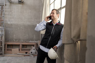 Smiling male contractor talking over smart phone while standing in building - VEGF03571