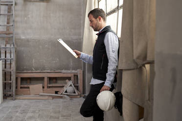Männlicher Bauunternehmer, der in einem Gebäude stehend ein Dokument analysiert - VEGF03572