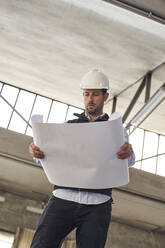 Männlicher Bauunternehmer analysiert einen Lageplan, während er im Gebäude steht - VEGF03573