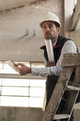 Male engineer with digital tablet and blueprint contemplating while standing in building at site - VEGF03584