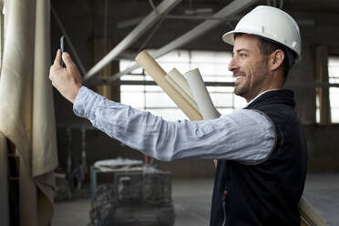 Lächelnder männlicher Architekt, der über sein Smartphone Videogespräche führt, während er in einem Gebäude steht - VEGF03589