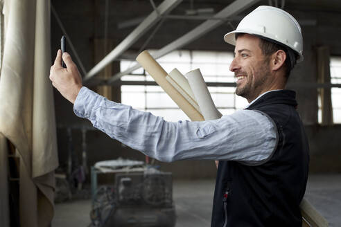 Lächelnder männlicher Architekt, der über sein Smartphone Videogespräche führt, während er in einem Gebäude steht - VEGF03589