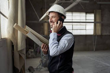 Smiling male building contractor talking over smart phone inside construction site - VEGF03590