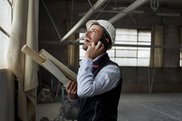 Cheerful male architect laughing while talking over mobile phone in constructing building - VEGF03591