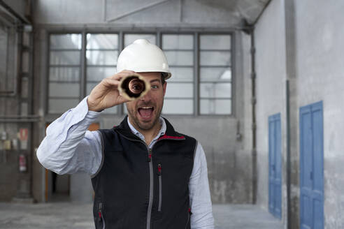 Männlicher Architekt mit offenem Mund, der durch einen Karton schaut, während er in einem Gebäude steht - VEGF03596
