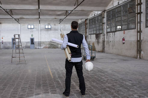 Männlicher Architekt hält Kartons und Schutzhelm, während er auf dem Boden eines Gebäudes steht - VEGF03602
