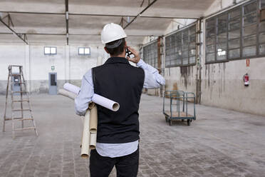 Male architect holding cardboards talking over mobile phone while standing in building - VEGF03603