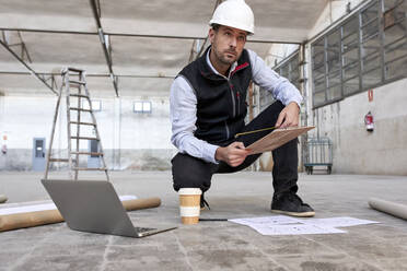 Männlicher Architekt, der einen Lageplan hält, während er in einem Gebäude auf dem Boden hockt - VEGF03608