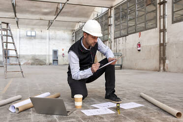 Männlicher Ingenieur analysiert einen Lageplan, während er in einem Gebäude auf dem Boden kniet - VEGF03610