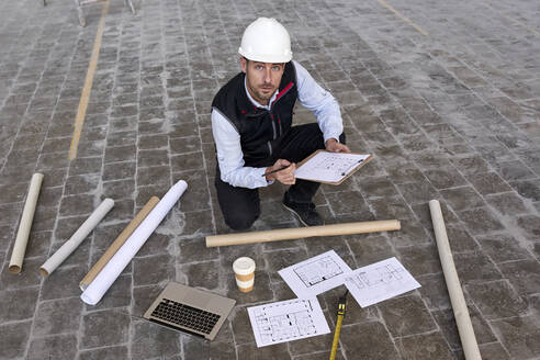 Männlicher Ingenieur hält einen Lageplan, während er in einem Gebäude auf dem Boden kniet - VEGF03612