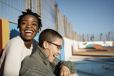 Smiling friends looking away while sitting together at sports court - RCPF00559